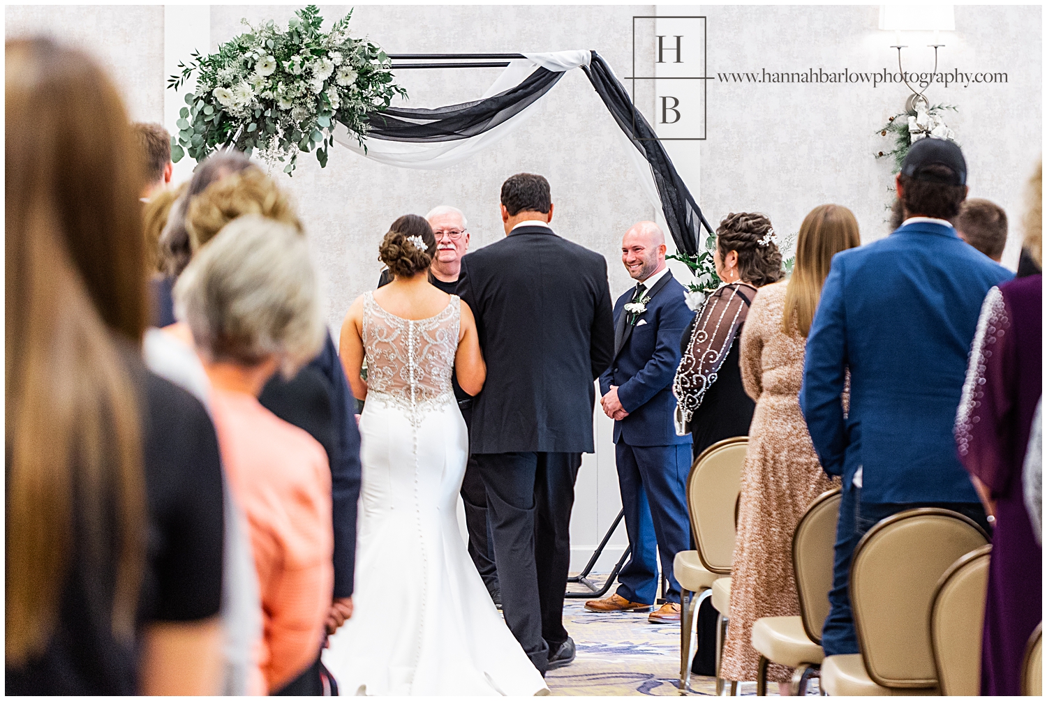 Photo from the back of wedding ceremony with groom looking at bride being escorted by father.