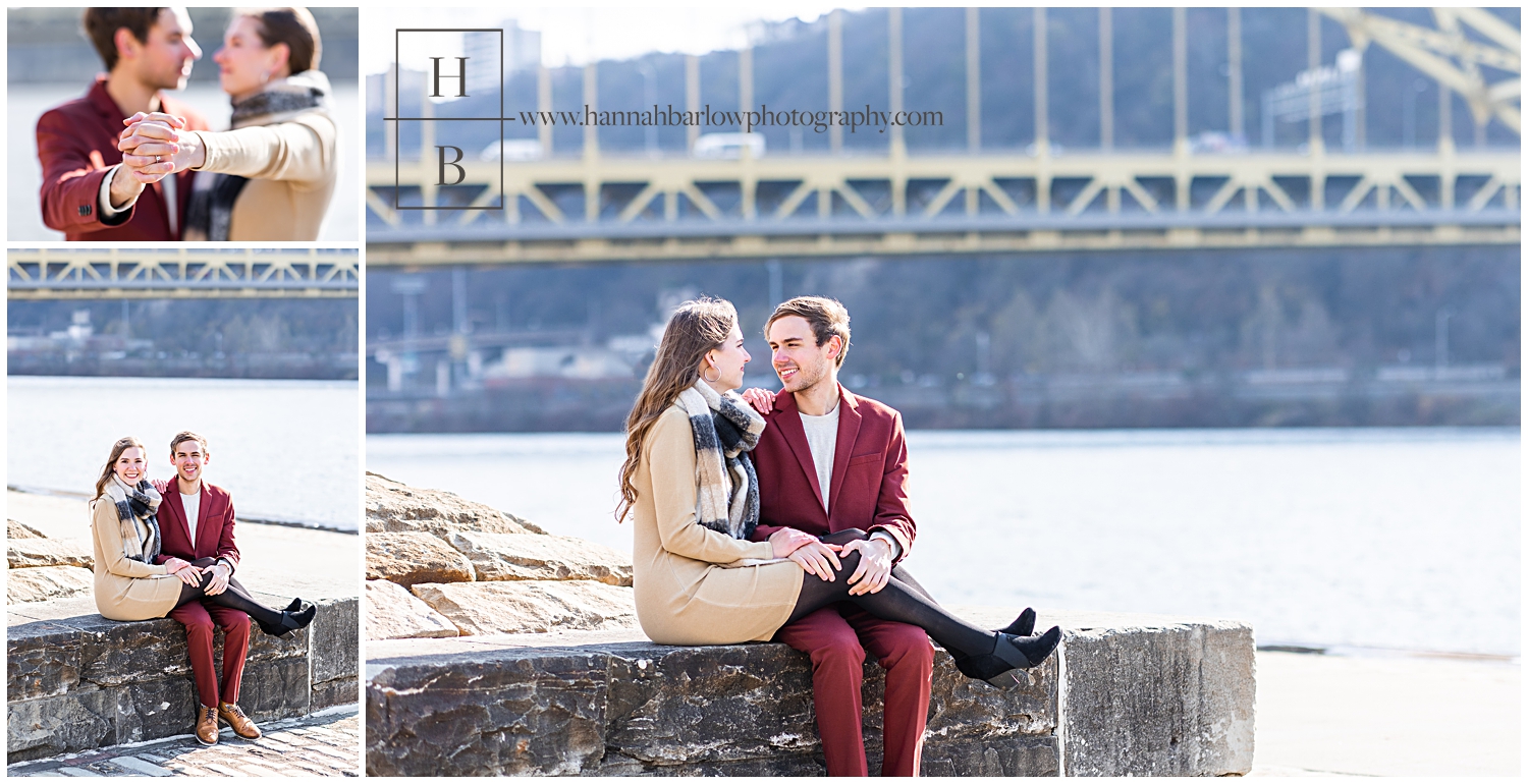 Couple sits on rock stone wall in front of Pittsburgh bridge.