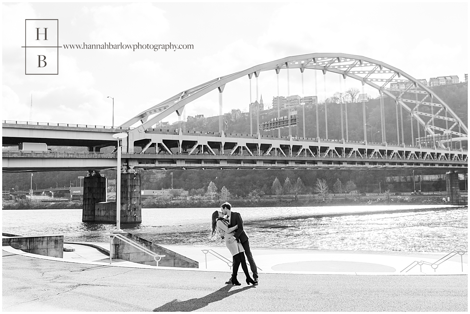 Black and white photo of couple dipping and kissing in front of Pittsburgh bridge.