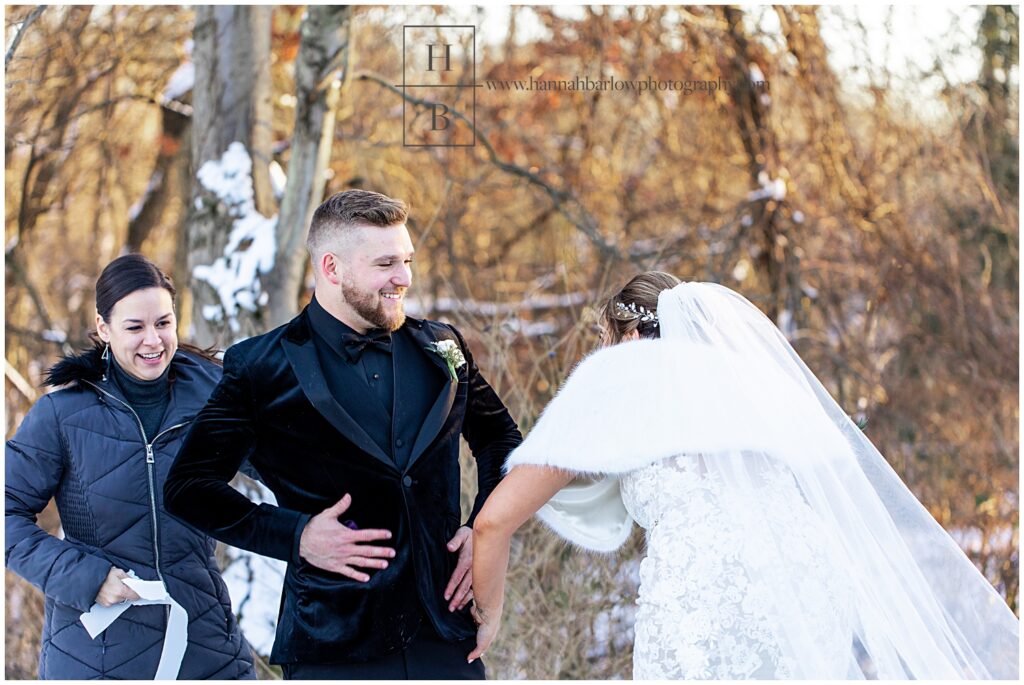 Groom in black velvet tux laughs next to bride and wedding photographer