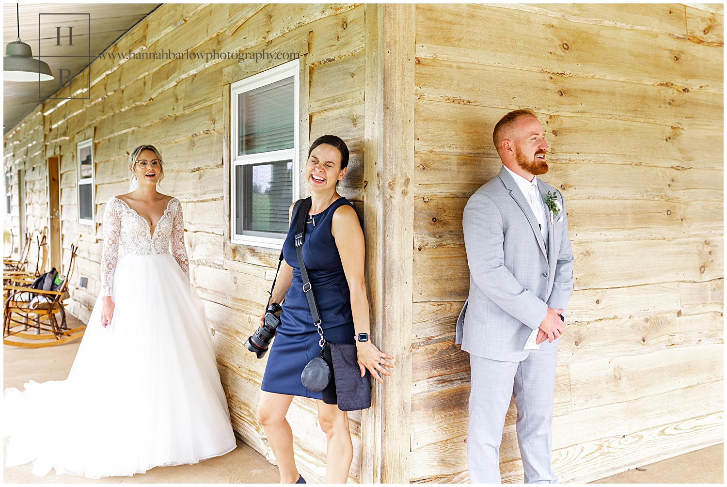 Wedding photographer stands with groom against barn and laughs.