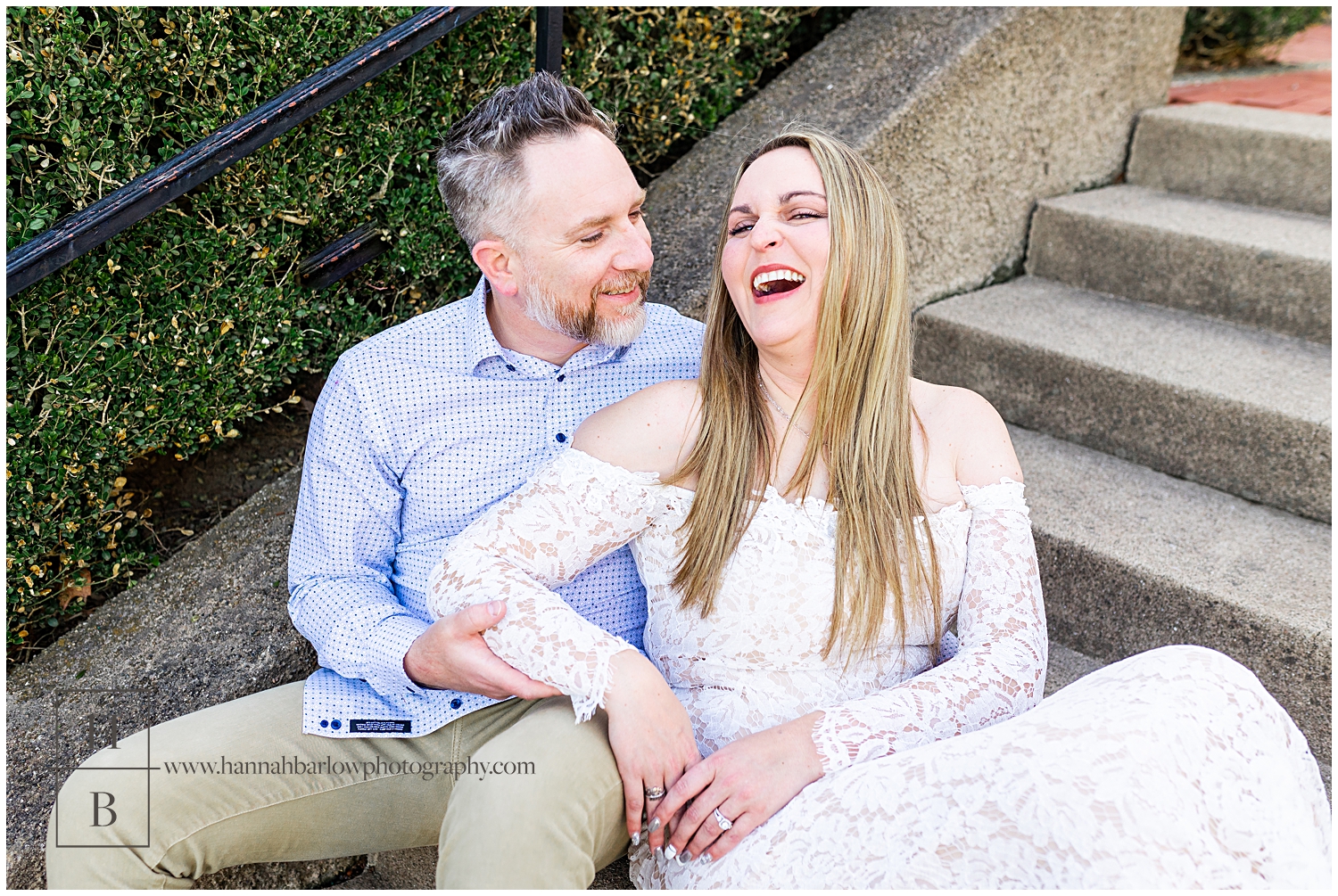 Girl in white lace dress laughs while being embraced by fiancé on stone steps.