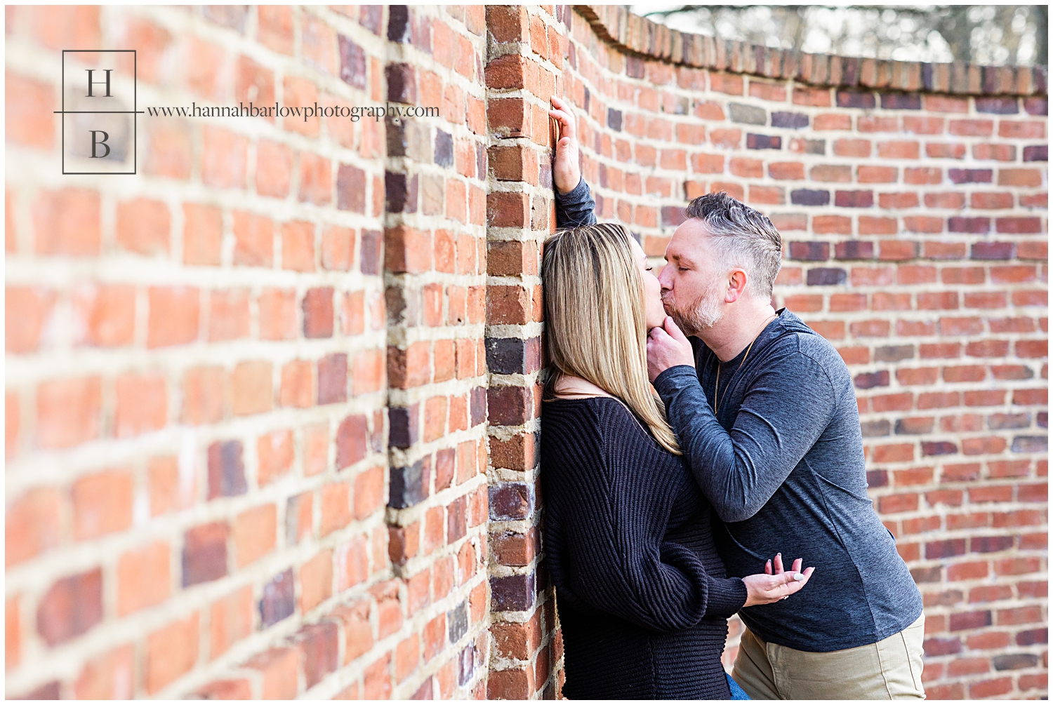 Groom to be embraces fiancé and brings chin in for a kiss against brick wall.