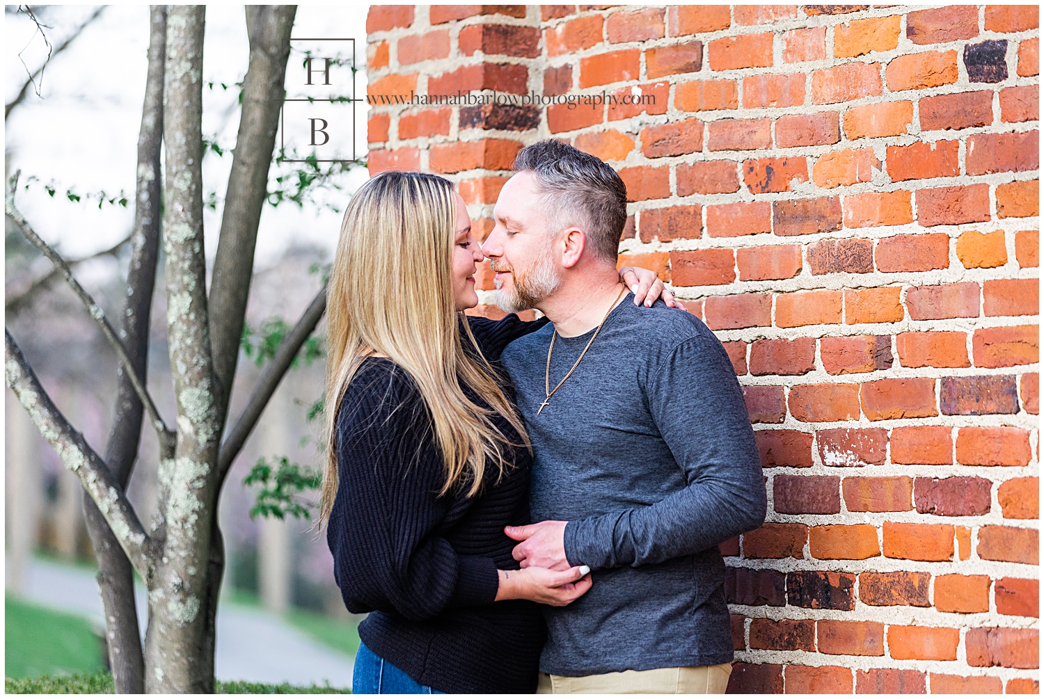 Couple stands nose to nose by brick wall for engagement photo.