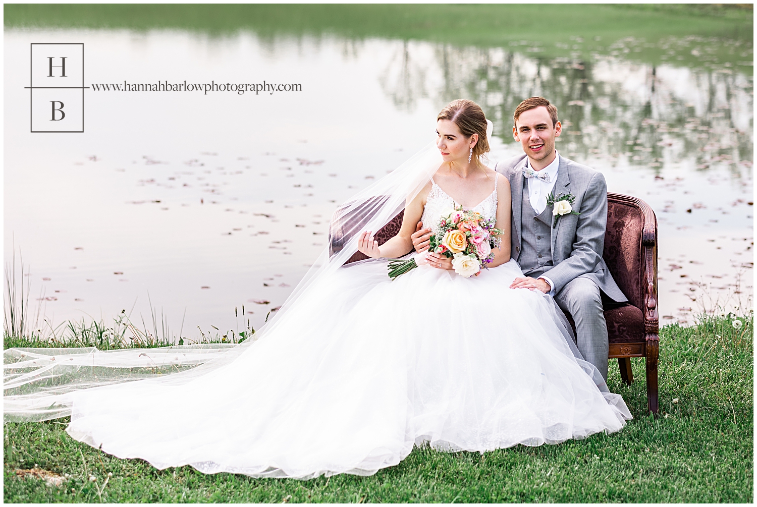 Bride and groom sit on chair by lake while bride looks on and groom looks at camera.