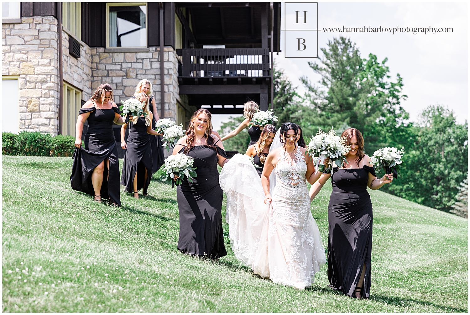 Bridesmaids in black dresses help bride walk down hill.