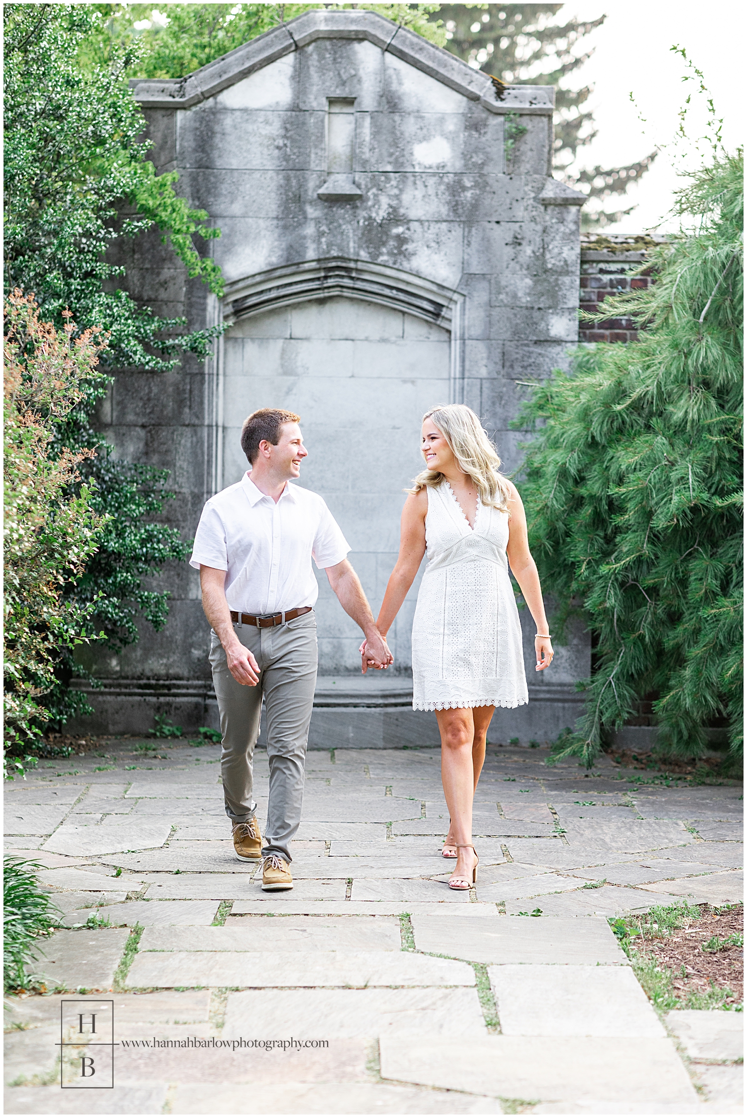 Couple walks by lush green foliage and stone wall for engagement photos.
