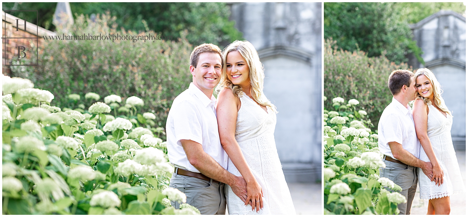 Lady in white dress snuggles up to fiance by white hydrangea bushes.