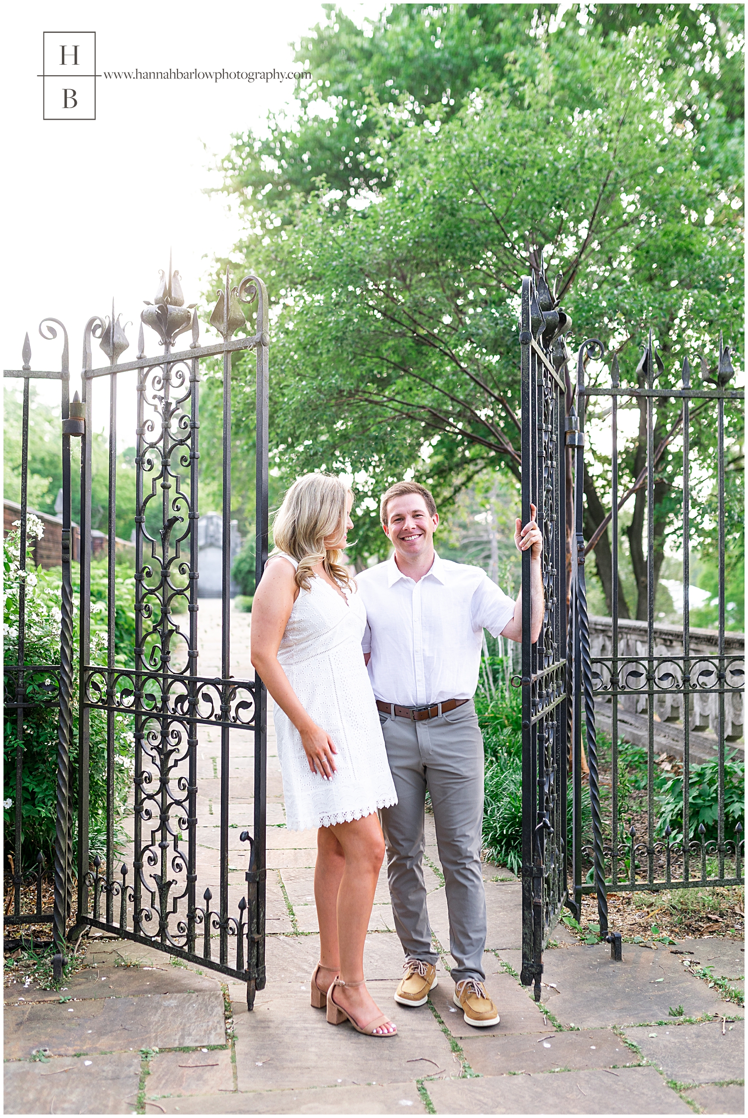 Groom stands in black iron gate opening holding embracing fiance for engagement photos.