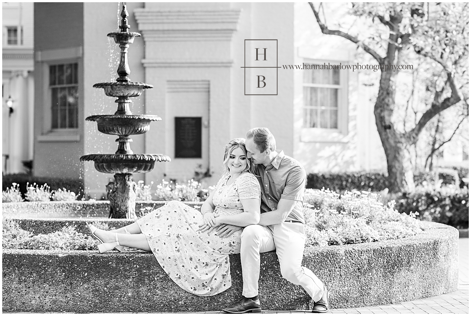 Man and fiancé pose on stone wall in front of water fountain and embrace for engagement photos.