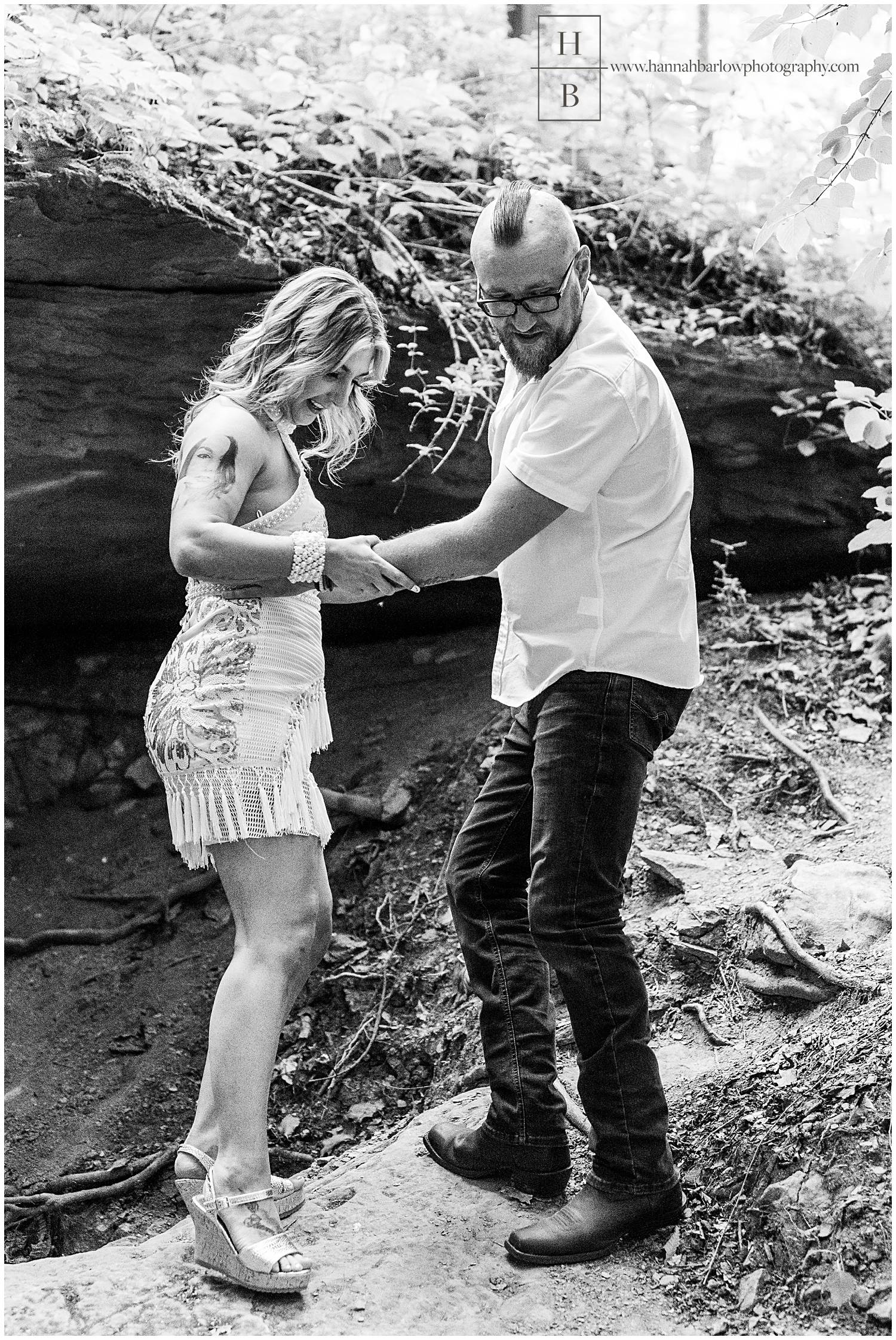 Black and white photo of man helping fiance down while hiking through rocks.