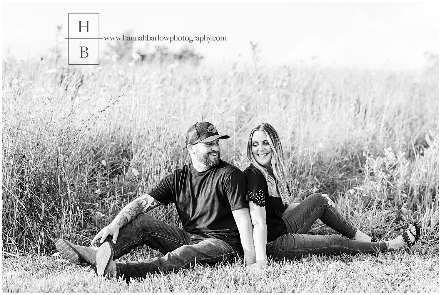 Black and white photo of couple sitting on field weed line looking back at one another for engagement photos.