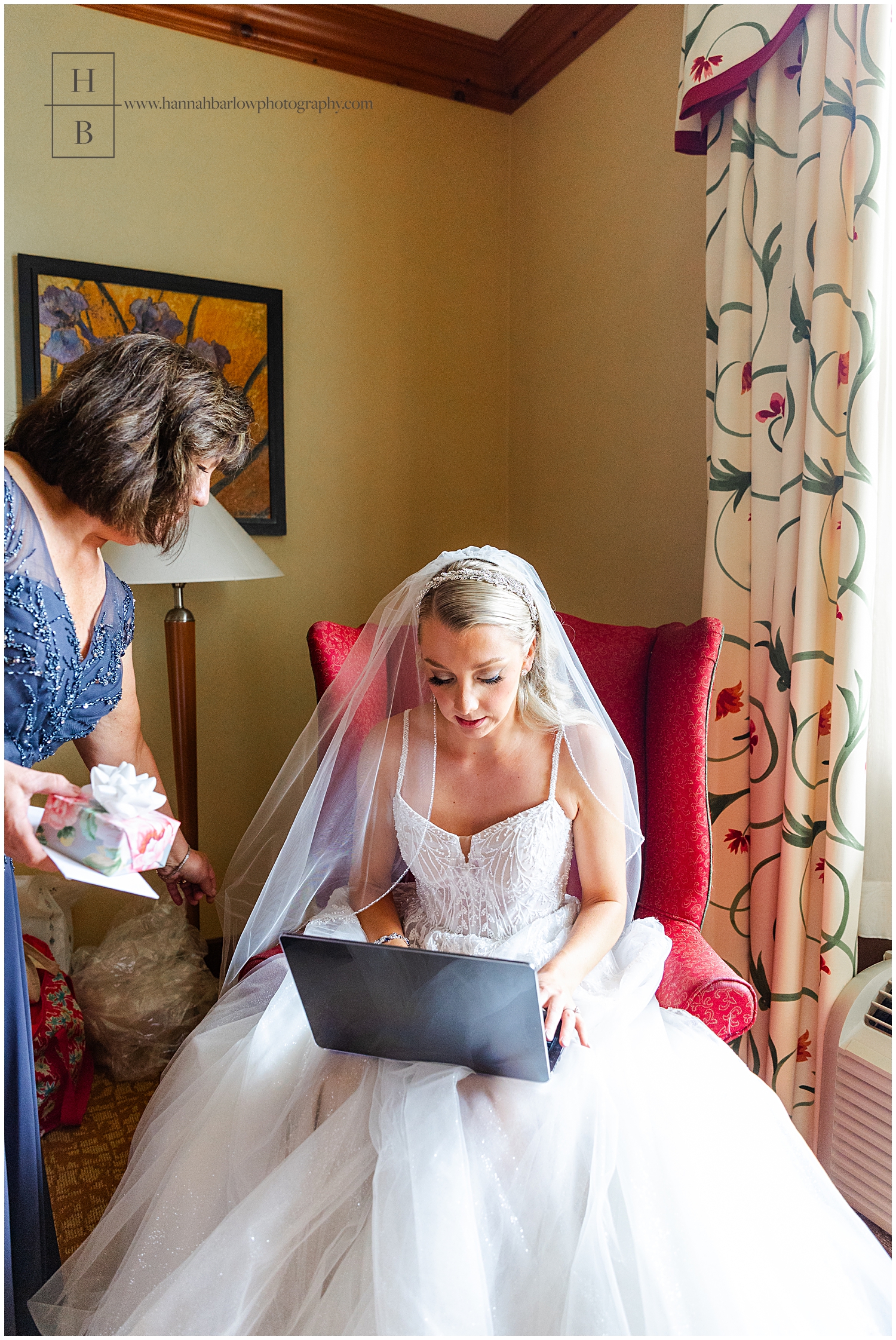 Bride holds computer on lap with mom standing at side.