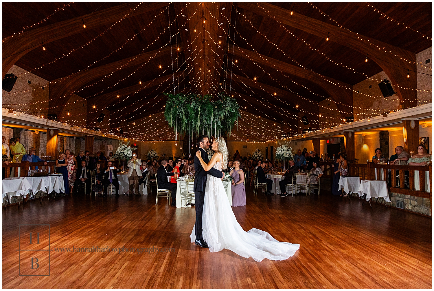 Bride and groom embrace for first dance with guests and tables in background under gold lights.