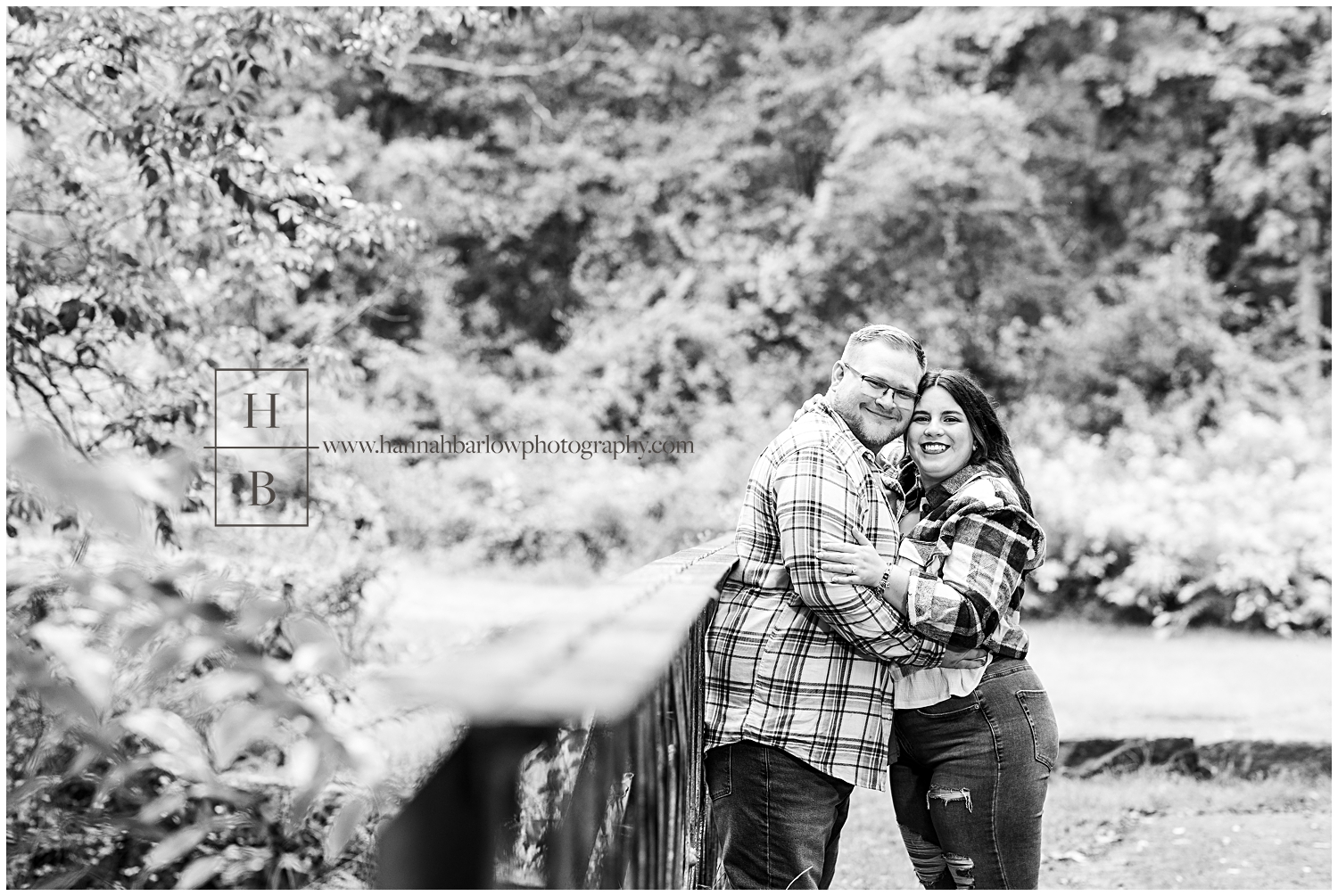 Black and white photo of couple standing against bridge embracing for photo.
