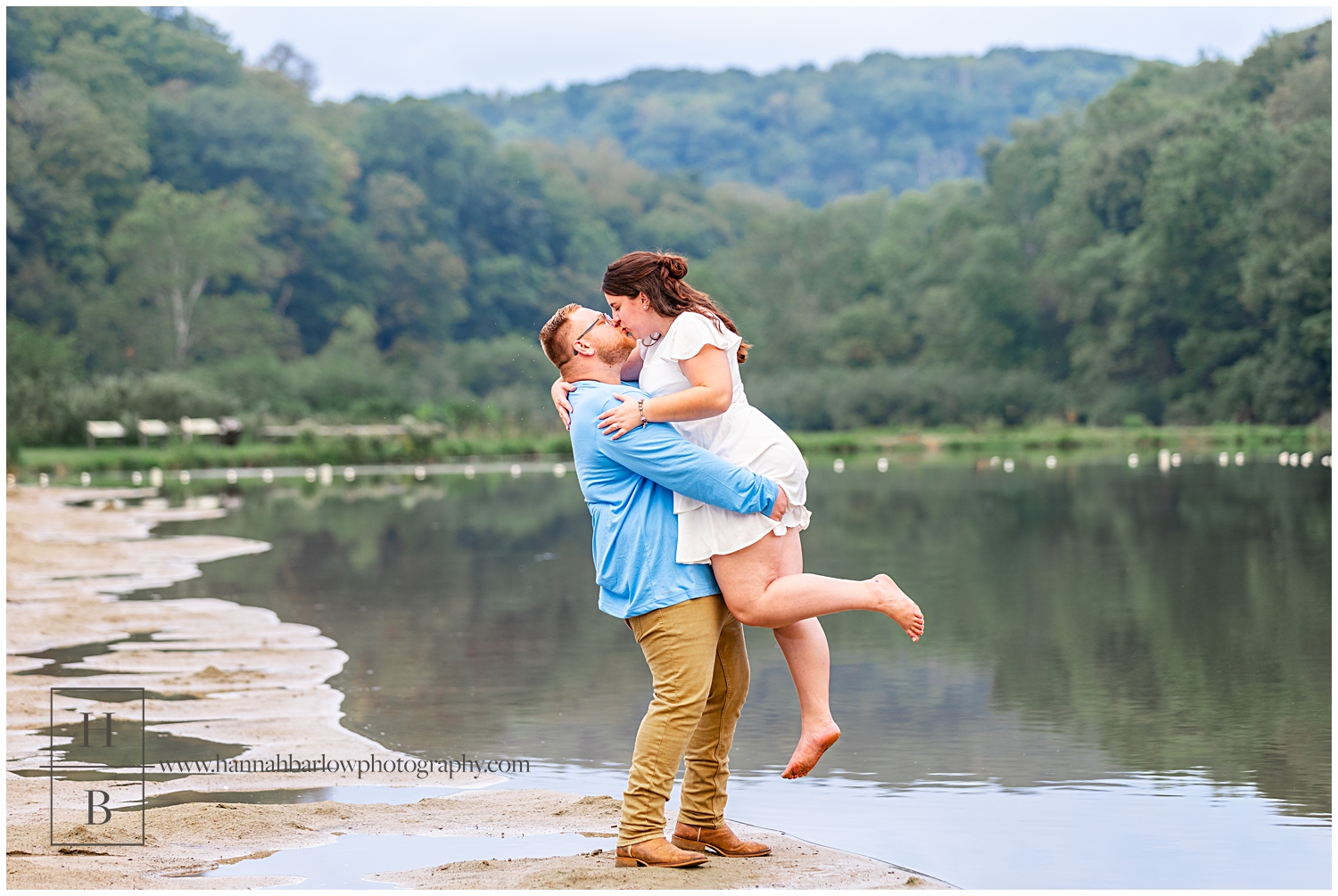 Man in blue shirt holds fiancé in white dress up in the air for a kiss.