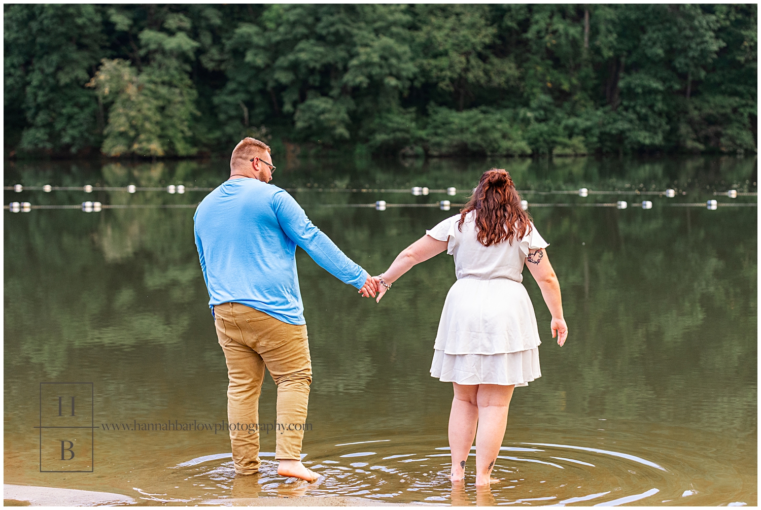 Couple holds hands and walks into lake water for engagement photos.