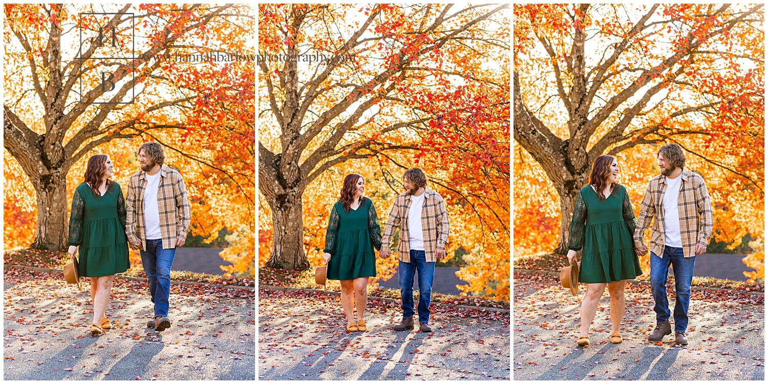 Couple holds hands and walks in front of trees with orange leaves.