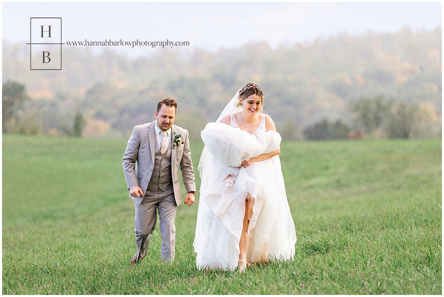 Bride carries dress through field as groom follows behind.