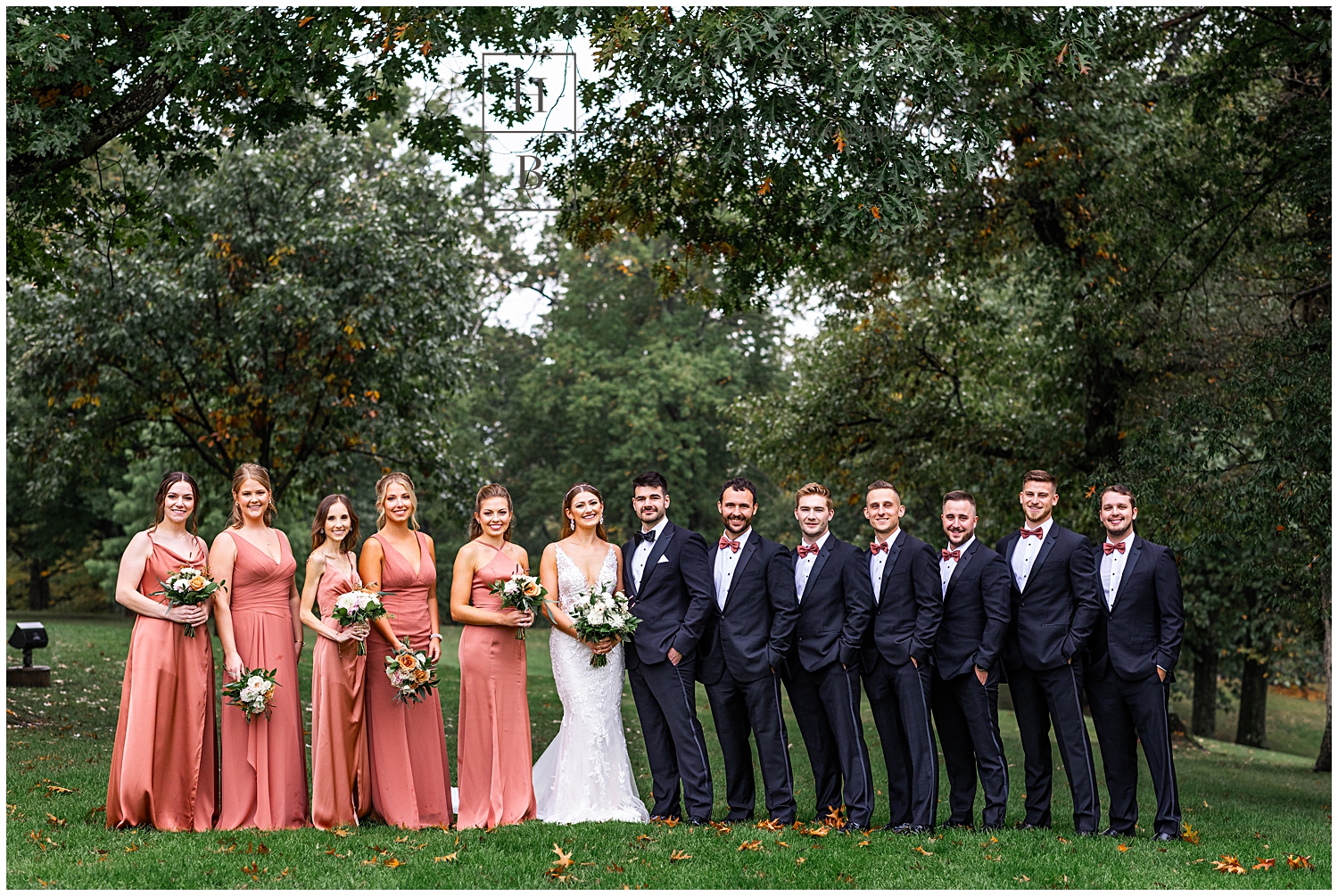 Bridesmaids in cedar rose dresses pose with groomsmen in black tuxes for bridal party photo.