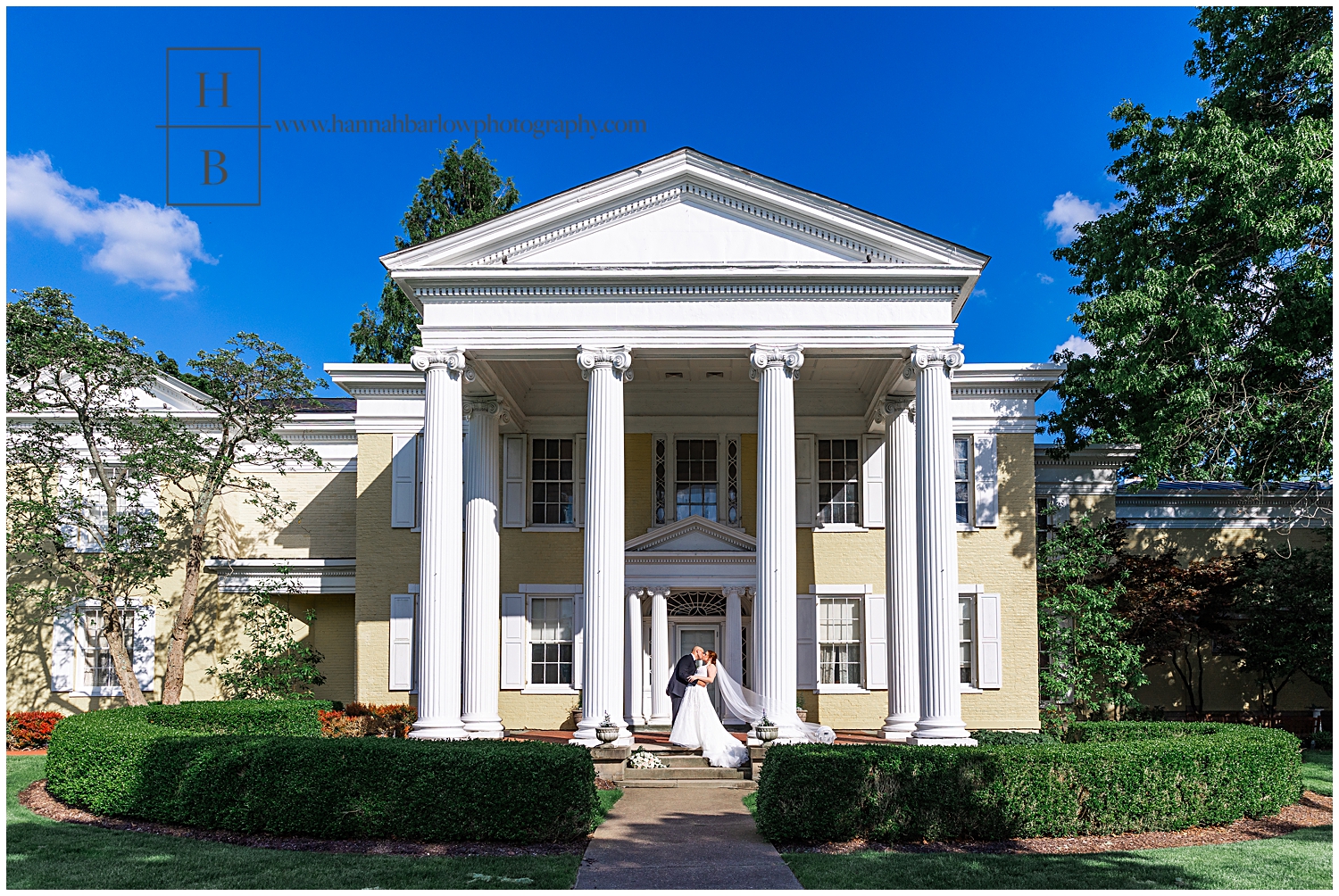 Bride and groom kiss in front of yellow mansion while veil flies in the wind