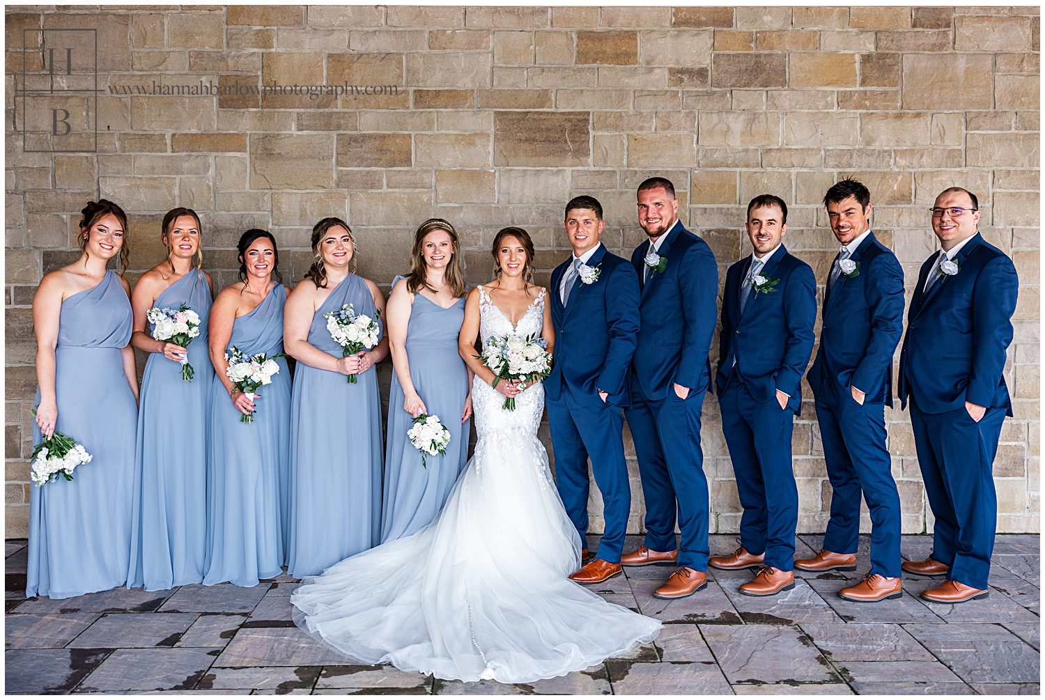 Bridesmaids in dusty blue dresses pose with groomsmen in navy tuxes with wedding couple