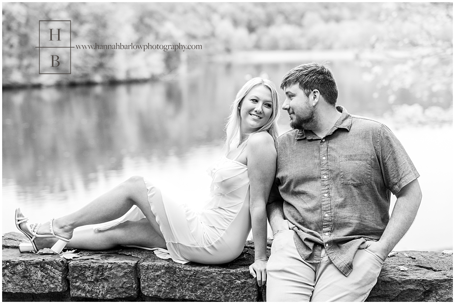 Black and white photo of woman sitting on brick wall embracing fiancé