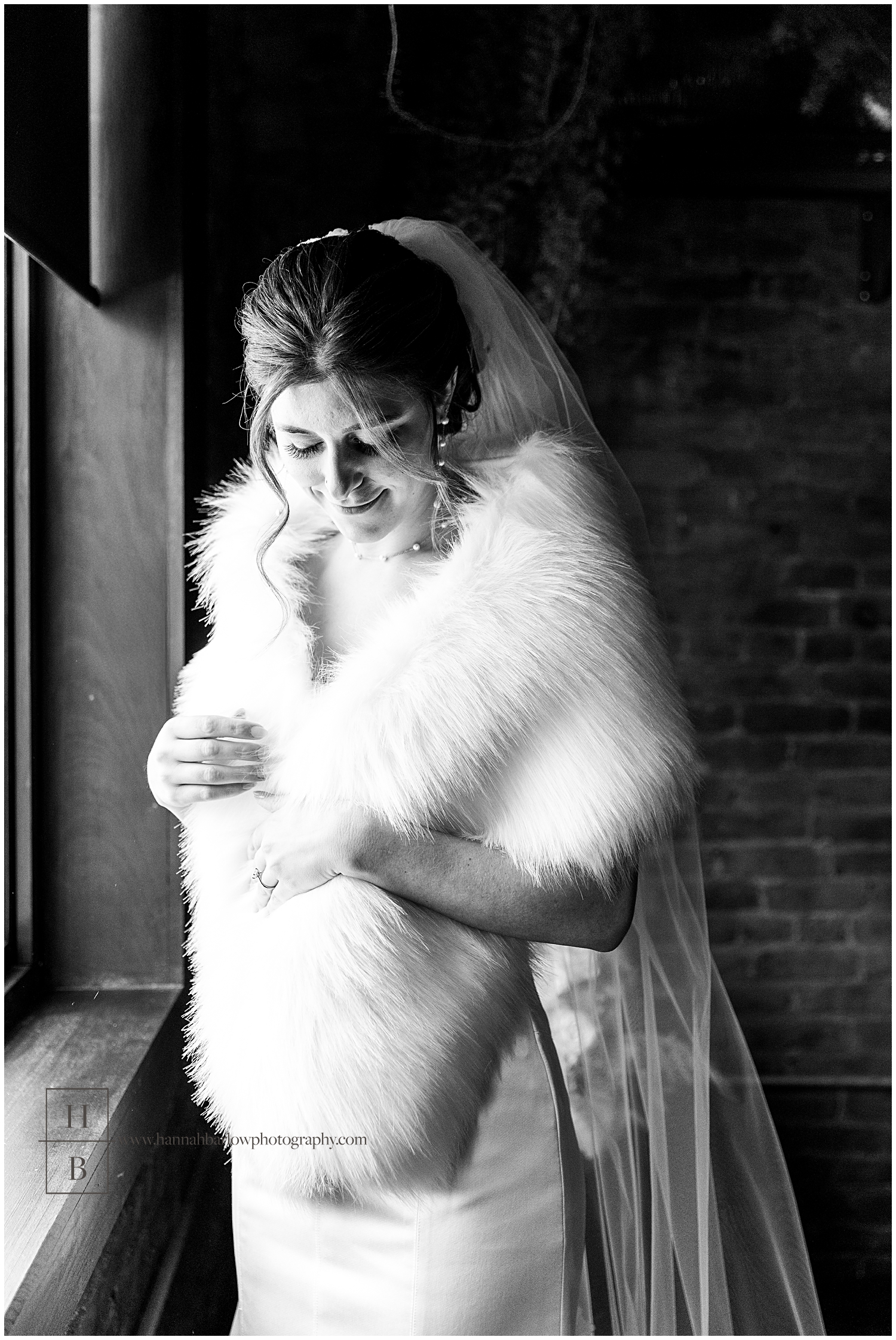 Black and white photo of bride standing by window wrapped in fur Shawl