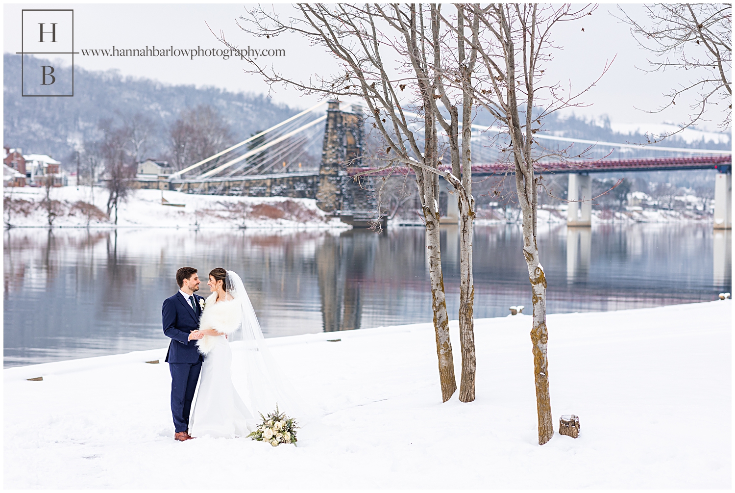 Bride and groom pose in snow with bridge in background