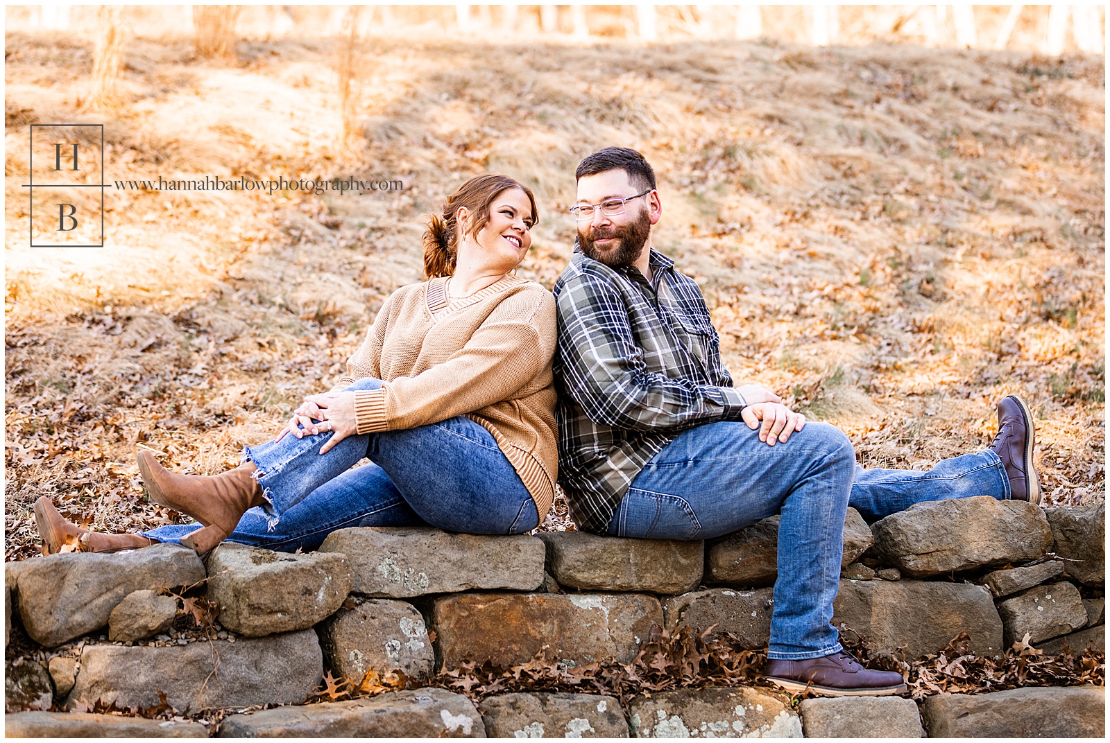 Couple sits on rock wall and looks at one another for engagement photos