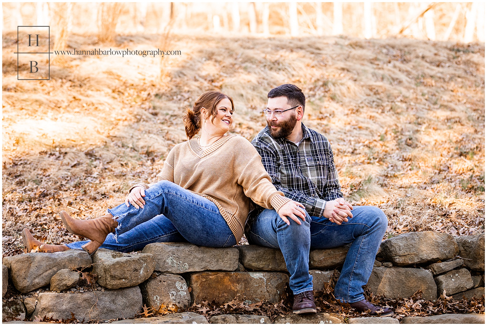 Couple sits on rock wall and looks at one another for engagement photos